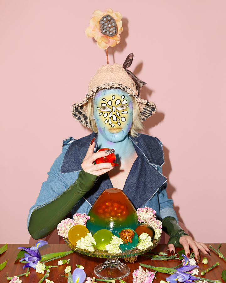 Jesse Clark posing behind a rainbow jello cake with patterned facepaint on against a pink background.
